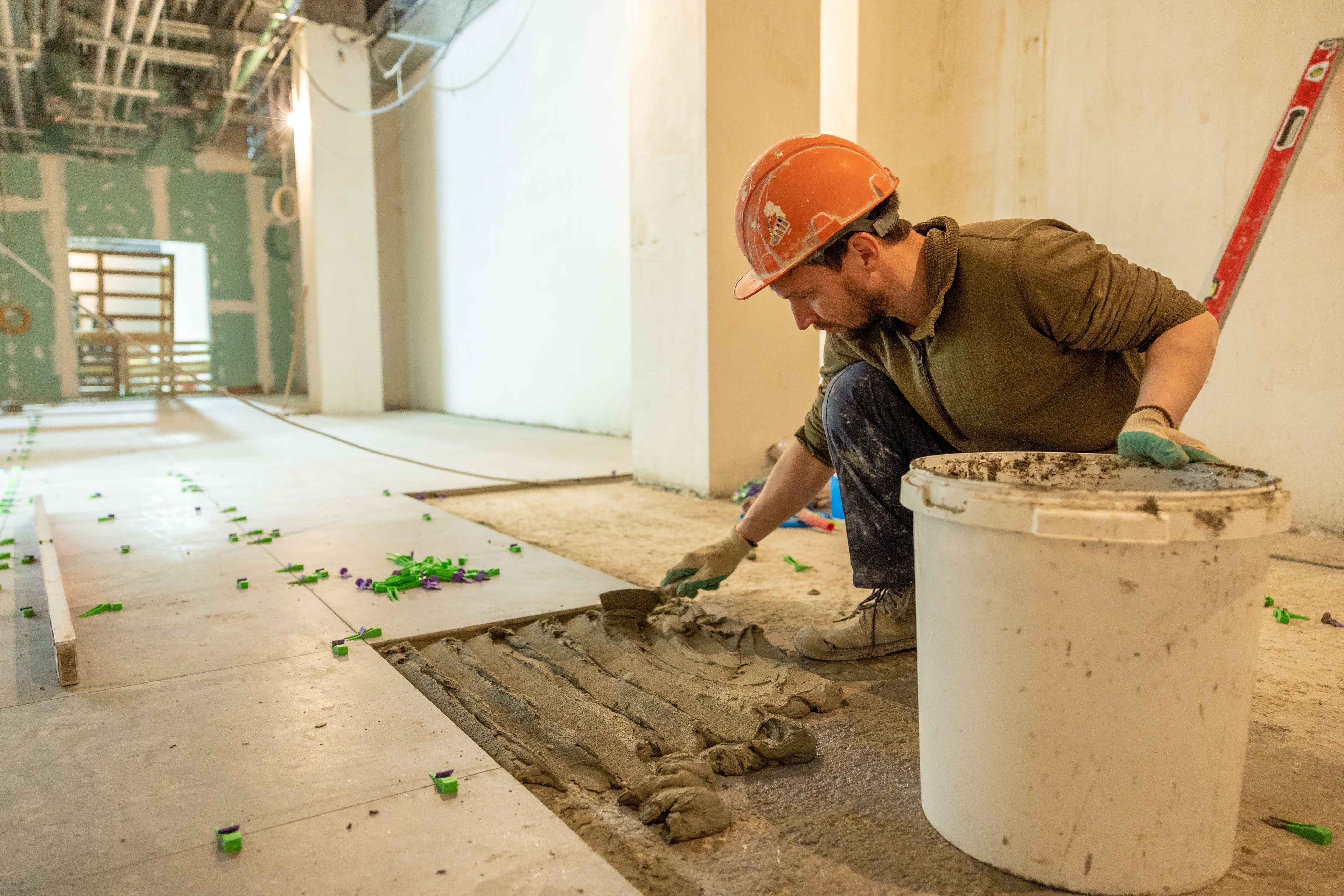 Un carreleur étale la colle au sol, un seau à ses côtés, sur un chantier en lumière du jour